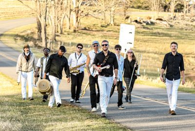 Nine musicians dressed in black and white, walk down a rural road carrying various musical instruments