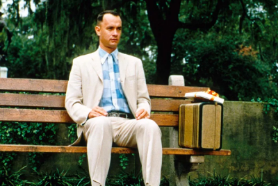 Forrest Gump (Tom Hanks) sits on a bench outdoors with a suitcase beside him.