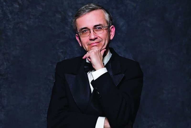 A man, smiling, wearing glasses, with his chin resting in his hand.