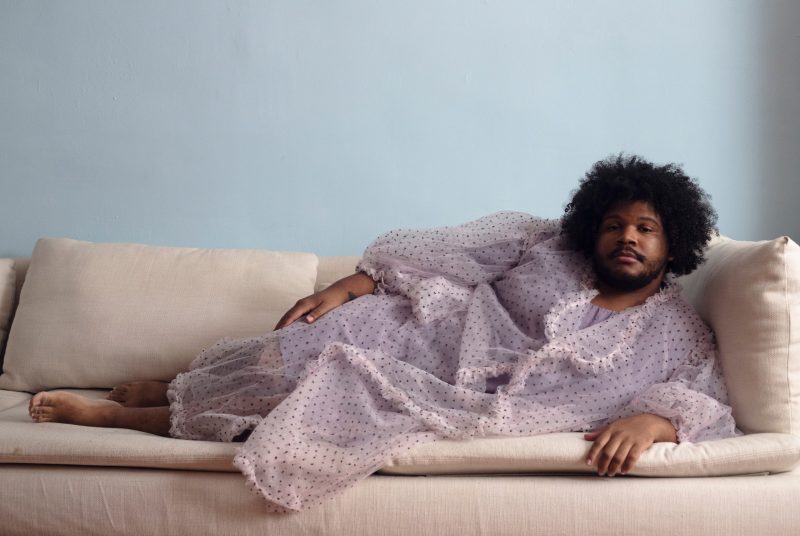 Bassoonist and composer Clifton Joseph Guidry III reclines on an off-white sofa, barefoot and dressed in a  light pink polka dot robe.