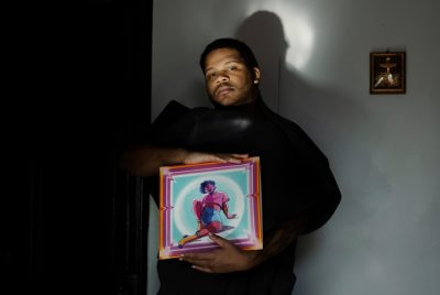 Joy Guidry, dressed in a long, black gown, holding a framed, colorful picture of a person in a pink shirt and blue shorts,sitting.