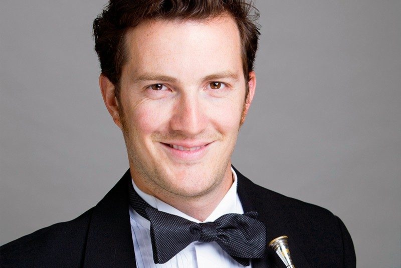 A man with short hair, smiling, in a tuxedo, holding a French horn