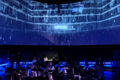 Musicians sit in the Cube black box theatre with catwalks above, in blue lighting