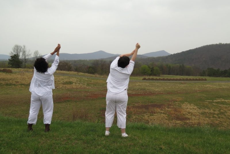 Two people dressed all in white stand outdoors in a field with the mountains in the distance, with their backs to the camera, arms outstretched overhead.
