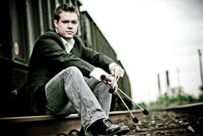 A young man wearing a dark jacket, white shirt, and jeans,  sits on a rail of the train tracks, holding two sets of percussion mallets in his hands.