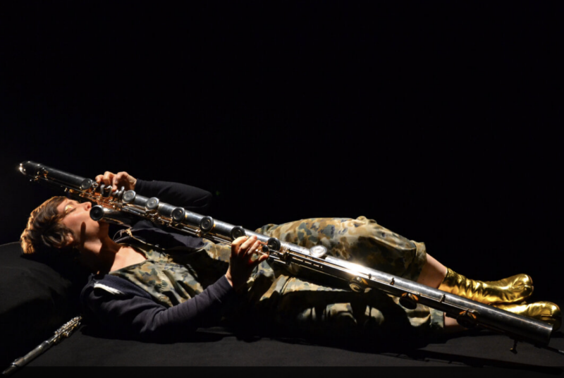 A woman wearing camo pants and top with a black jacket, reclining and holding a bass flute.