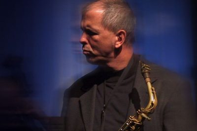 A man dressed in a black shirt and black jacket, holding a saxophone, with his head turned to the side.