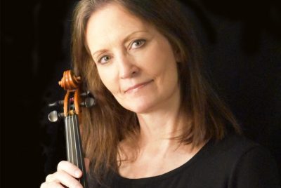 Donna Fairbanks, wearing a black top, with auburn mid-length hair, holding a violin.
