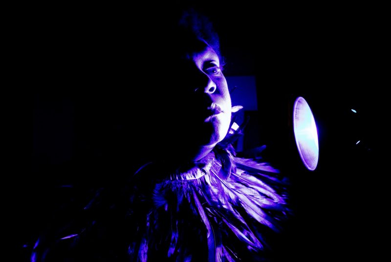 A photo of a woman's face, lit on one side with a purple light. She is wearing a necklace of feathers.