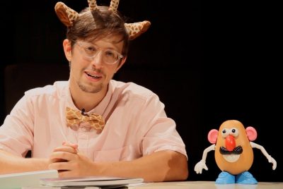 A young man with glasses,  wearing a short sleeved shirt, a bow tie in a giraffe print, and wearing glasses and giraffe horns on his head, with a Mr. Potato Head doll standing on the table in front of him