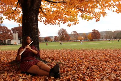 A young woman sits beneath a tree with orange autumn leaves on the tree and ground, playing a flute.