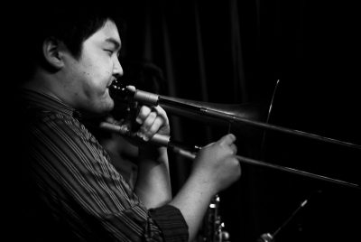 A black and white image of a dark haired man playing a trombone.