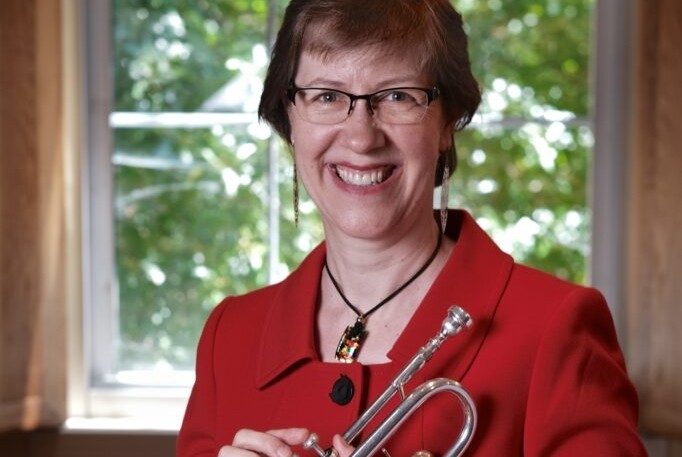 A woman with short hair and glasses, wearing a red top, smiling and holding a trumpet.