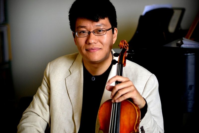 A dark haired man, with glasses, wearing a black shirt and off-white jacket, holds a violin and sits in front of a piano