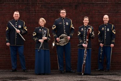 Five musicians of the US Army Woodwind Quintet stand in uniform holding their instruments.