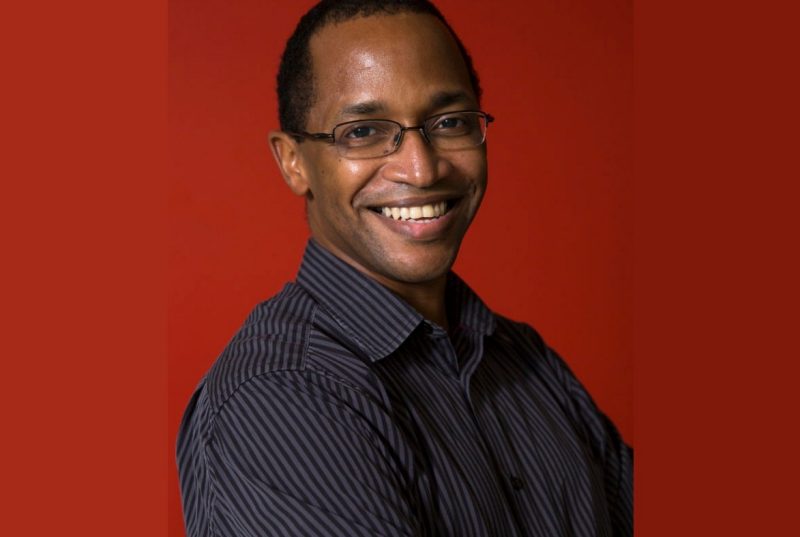 A black man, smiling, with short hair, wearing glasses and a grey and black striped shirt.