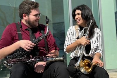 Hayden Scott, wearing a red shirt and holding two saxophones, sitting beside Layla Wilson, holding two trumpets