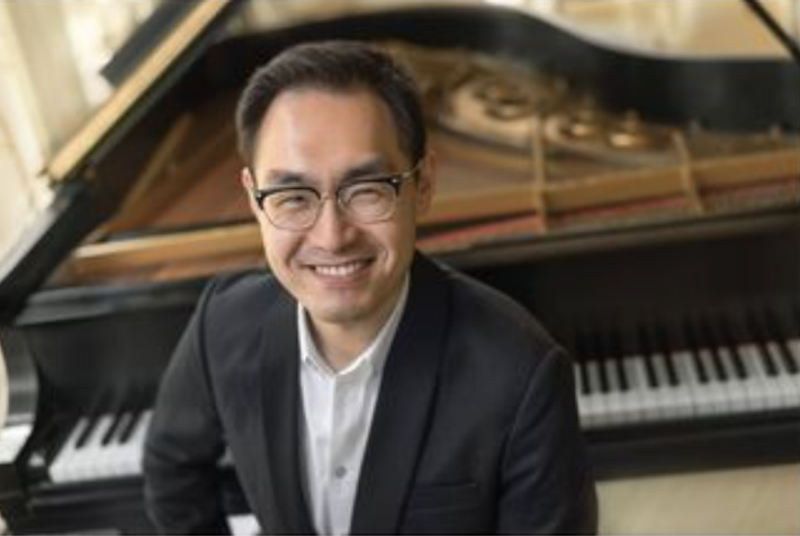 A man wearing a white shirt and black jacket, with glasses and dark hair, sits smiling in front of a grand piano.