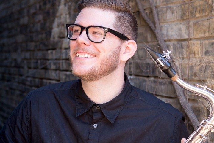 A man wearing a black shirt, and glasses, smiles, holding a saxophone