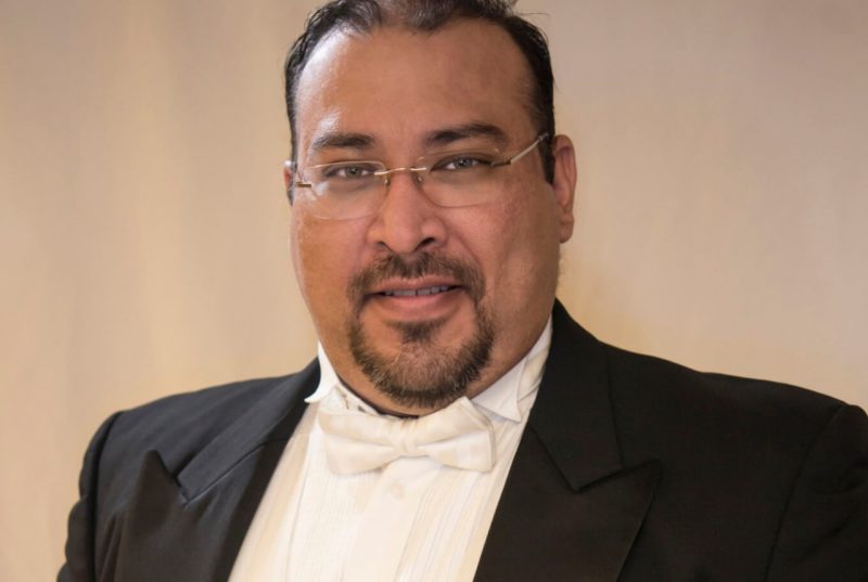 A man with dark hair, glasses, and wearing a tuxedo