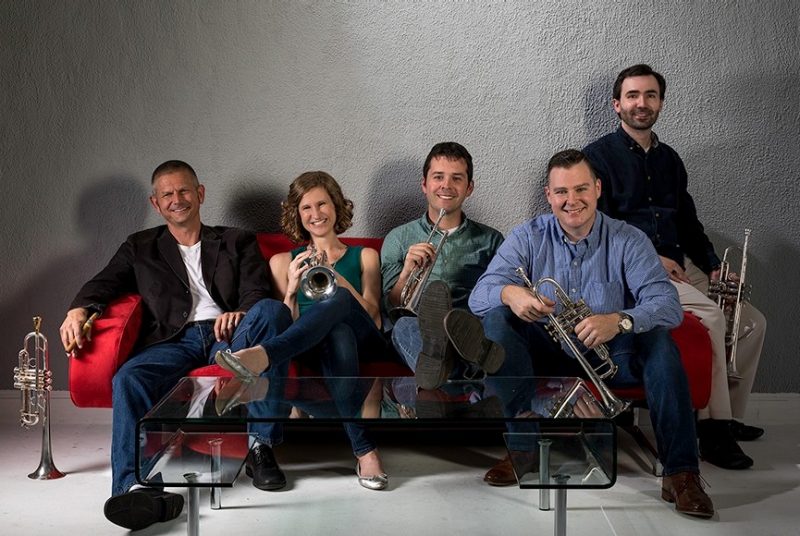 Three men and a woman sit on a red sofa, with another man standing behind, all with trumpets.