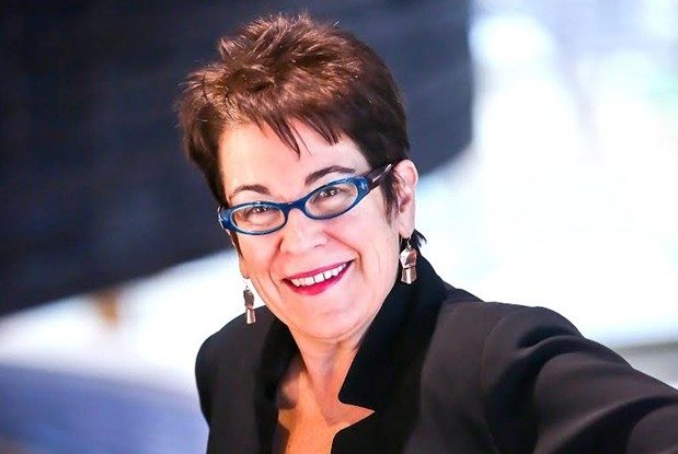 A woman with short reddish-brown hair ,wearing a dark top, red lipstick, blue-framed glasses, and earrings.
