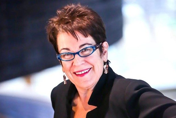 molly smith A woman with short reddish-brown hair ,wearing a dark top, red lipstick, blue-framed glasses, and earrings.