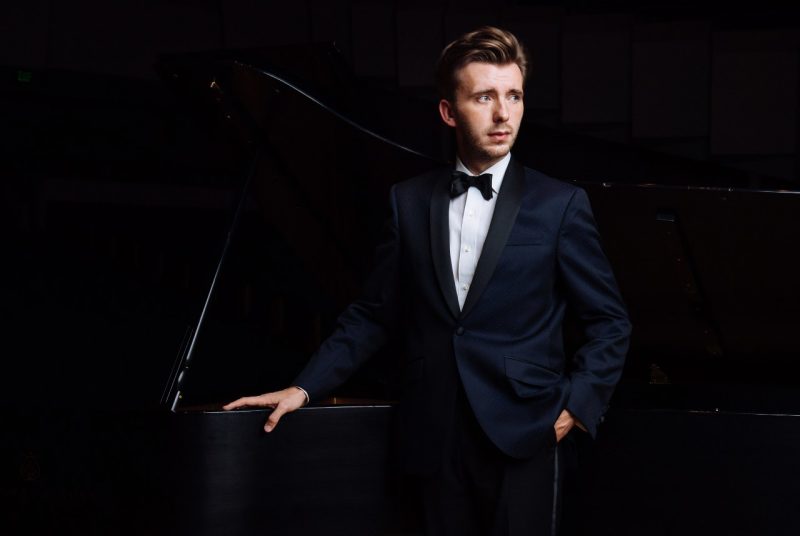 A man dressed in a tuxedo, brown hair, standing beside a grand piano.