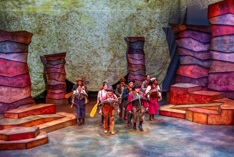 Ten actors on stage in stylized 'boats', some holding oars, with a set representing the Grand Canyon