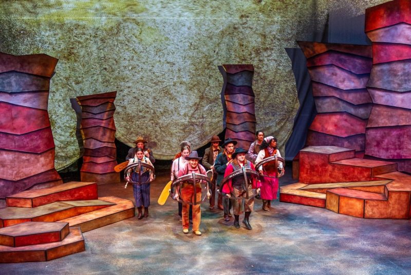 Men On Boats Ten actors on stage in stylized 'boats', some holding oars, with a set representing the Grand Canyon
