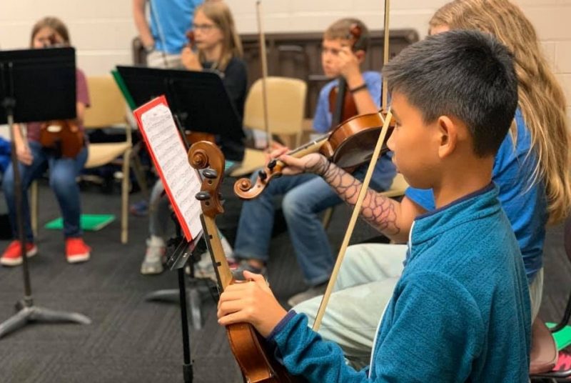 Five children sit with their stringed instruments in front of music stands with music.