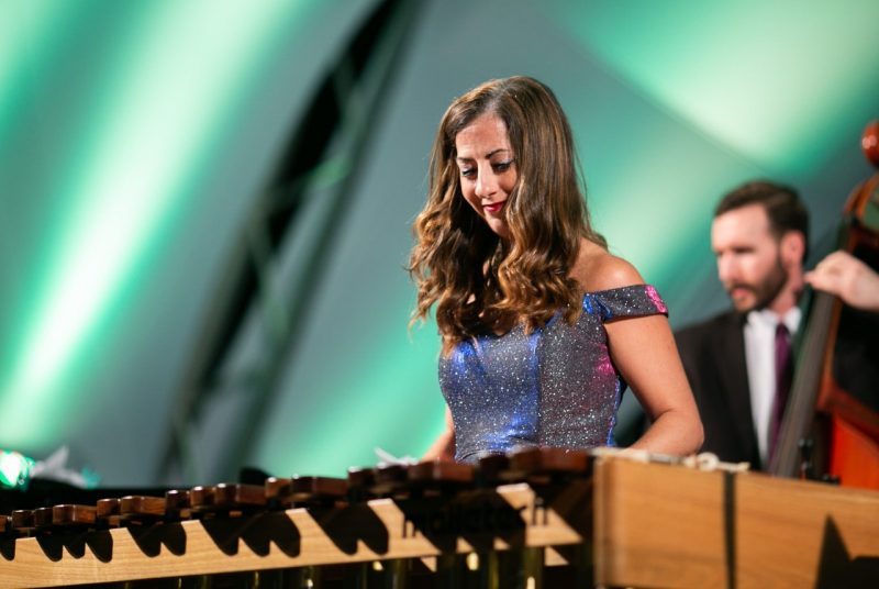 A woman with long hair plays a xylophone with a string bass player standing in the background.