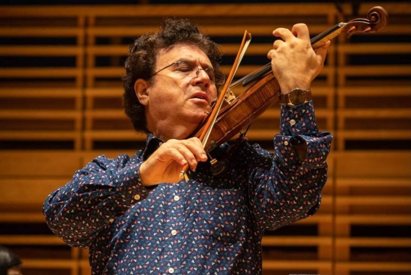 A man wearing glasses and a blue long-sleeved shirt plays a violin.