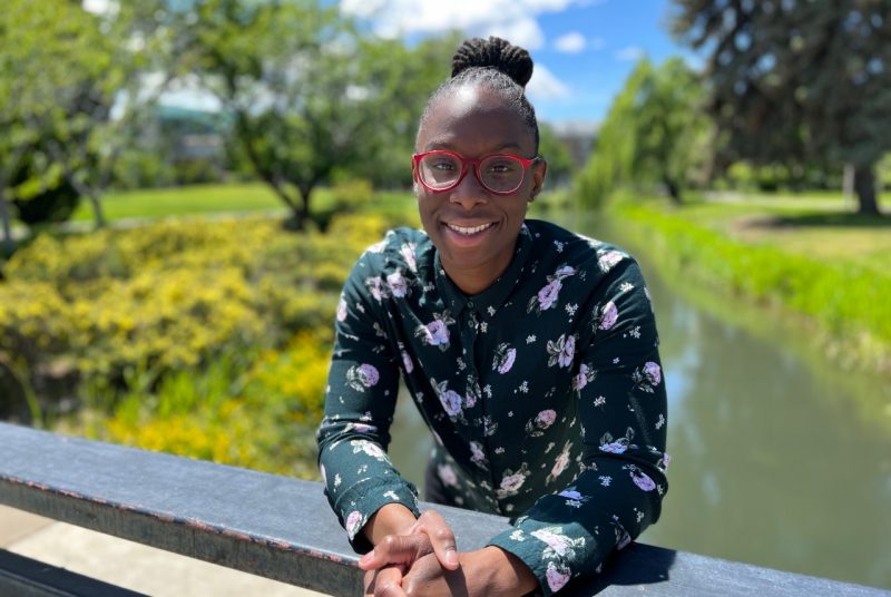 Kentra Wheeler A woman in a floral patterned long sleeve blouse, wearing glasses, stands on a small bridge that crosses a stream, with grass and trees in the background.