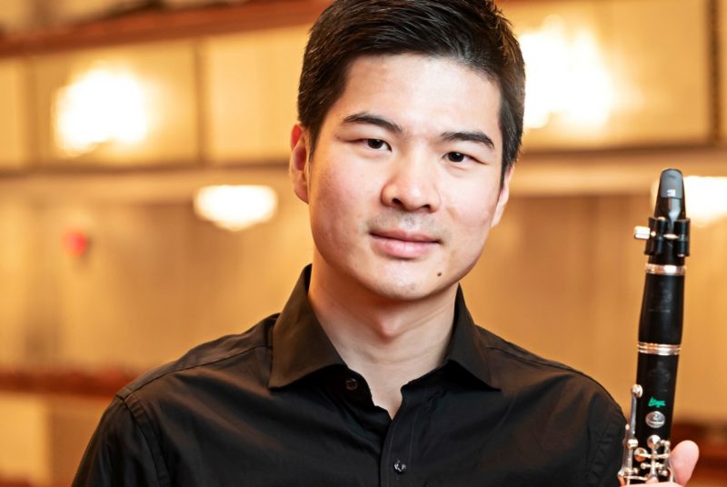 A man with short dark hair, wearing a black shirt, holds a clarinet.