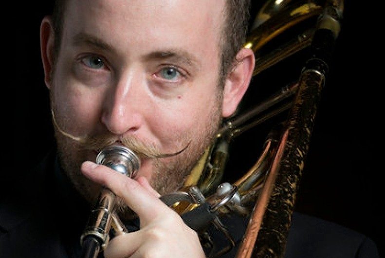 A man with short dark hair, wearing a black shirt, holds a clarinet.